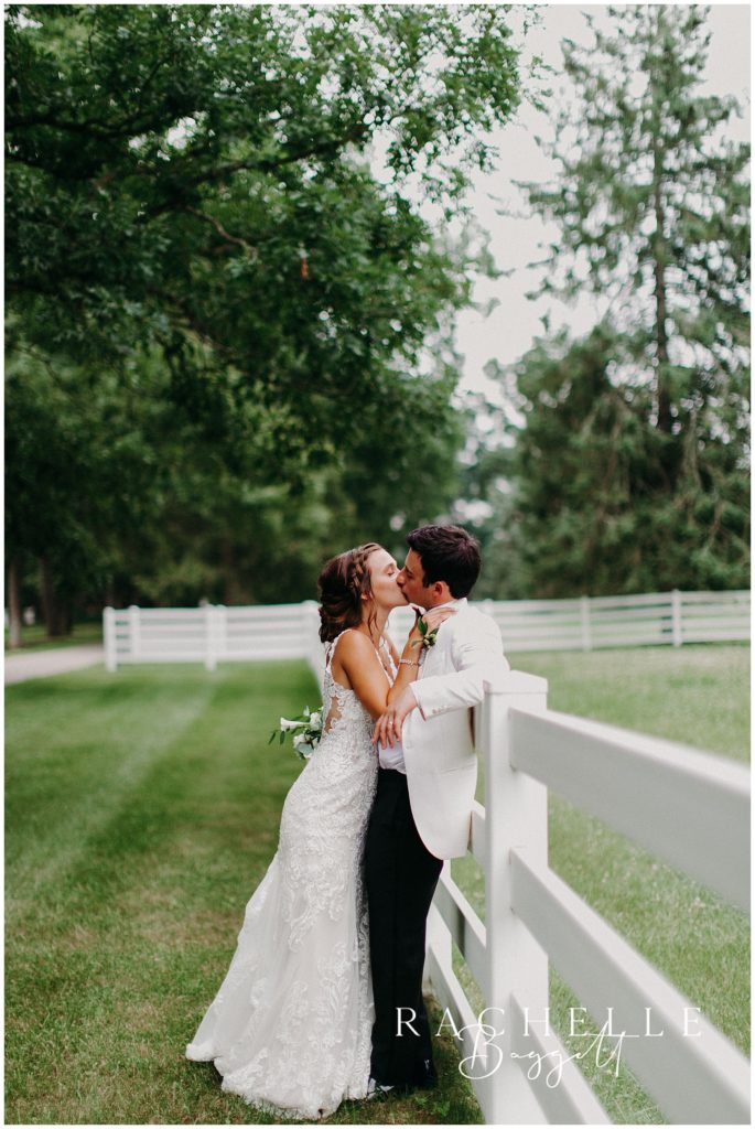 a bride and groom kiss and lean against a fence at camp tamarack in brighton, michigan
