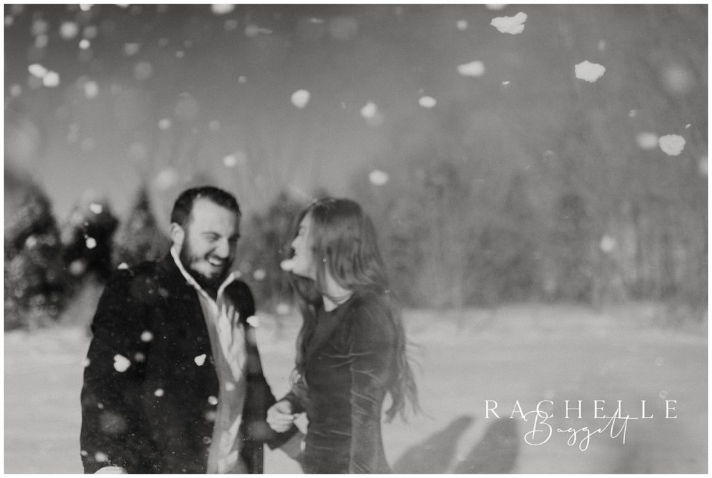 black and white photo of man and woman laughing while snow falls around them during their snowy sunrise engagement session