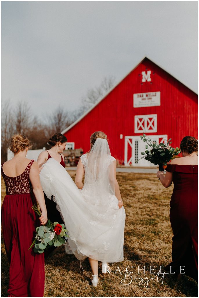 bridesmaids walking in front of red barn, helping bride with dress