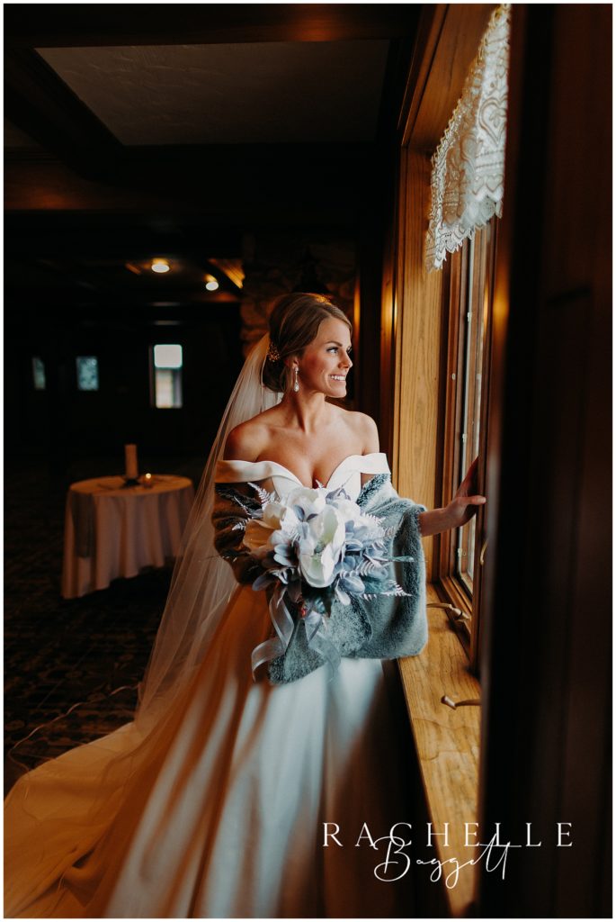 bride gazes out window with flowers in hand