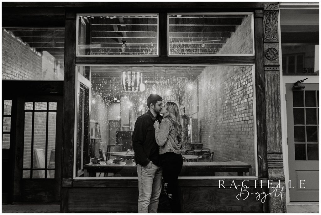 engaged couple kiss in front of glass window