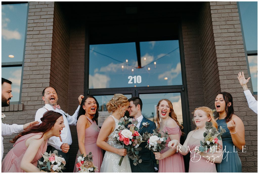 husband and wife lean in for kiss with bridal party cheering - Spring Wedding at City View